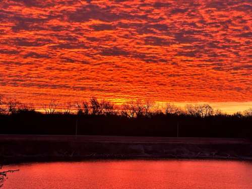 Vibrant sunset with fiery orange and red clouds reflecting on a calm body of water, surrounded by silhouettes of trees.
