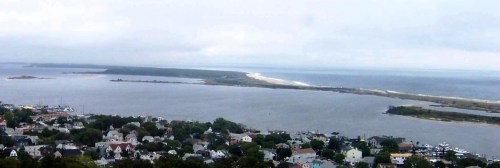 A coastal view featuring a river, sandy shore, and distant land under a cloudy sky, with houses in the foreground.