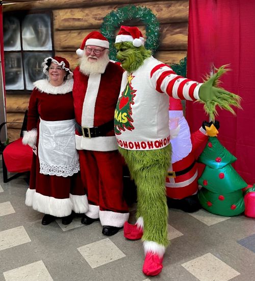 Santa, Mrs. Claus, and the Grinch pose together in festive attire, surrounded by Christmas decorations.