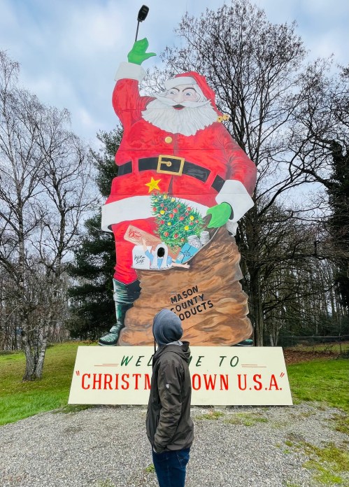 A person stands in front of a large Santa Claus statue at Christmas Town U.S.A., with trees in the background.