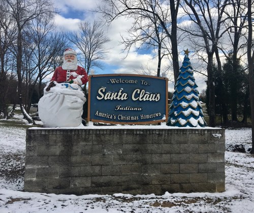 Welcome sign for Santa Claus, Indiana, featuring Santa and a Christmas tree, surrounded by snow and trees.