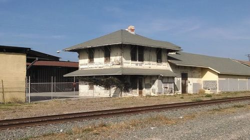 Abandoned two-story building with a sloped roof, near railway tracks, surrounded by overgrown grass and a fence.