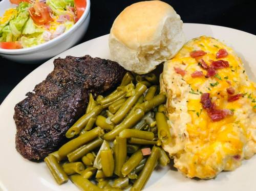 A plate with grilled steak, green beans, cheesy casserole, a biscuit, and a side salad.