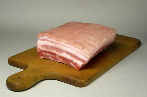 A piece of raw pork belly on a wooden cutting board, showcasing layers of fat and meat.