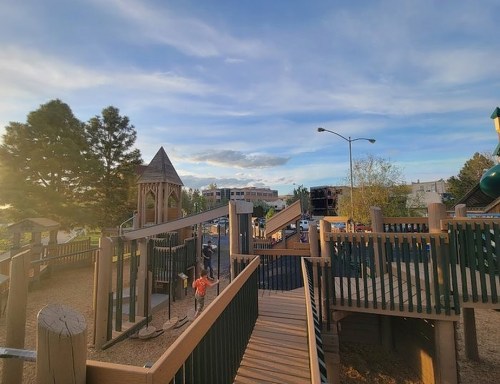 A playground with wooden structures, slides, and swings under a blue sky with scattered clouds. Children are playing.