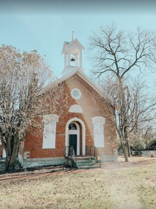 A brick church with a bell tower, surrounded by trees and a grassy area under a clear blue sky.