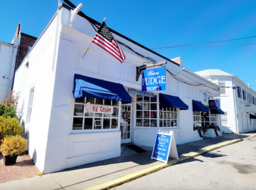 A white storefront with blue awnings, featuring a sign for "Fudge Shop" and an American flag.