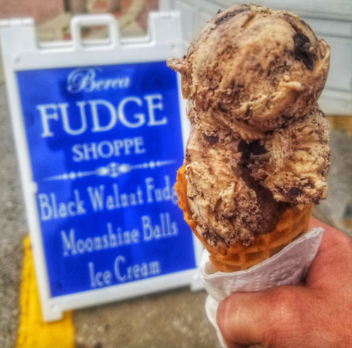 A hand holds a cone topped with chocolate and black walnut fudge ice cream in front of a sign for Berea Fudge Shoppe.