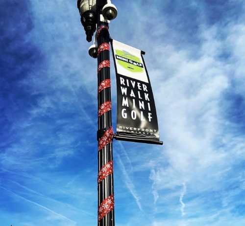 A street lamp with a banner for River Walk Mini Golf against a blue sky with wispy clouds.