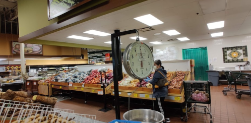 A grocery store interior with a scale in the foreground and a shopper browsing colorful produce in the background.