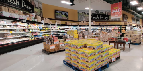 A grocery store aisle featuring dairy products, stacked boxes, and a sale sign, with shelves filled with various items.
