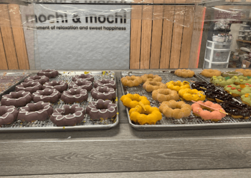 Trays of colorful mochi donuts in various shapes and flavors displayed in a shop.