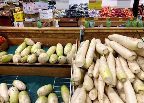 A display of fresh daikon radishes in a grocery store, with various fruits and vegetables in the background.