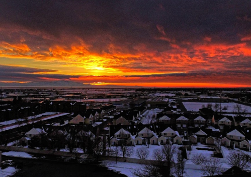 A vibrant sunset casts orange and purple hues over a snowy neighborhood and distant buildings.