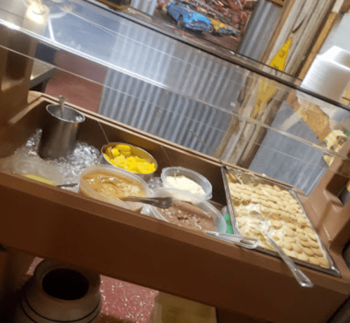 A dessert station with bowls of ice cream, toppings, and fruits displayed under a glass cover.