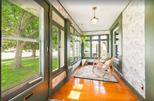 Bright sunroom with large windows, wooden floors, and a cozy rocking chair, surrounded by greenery outside.
