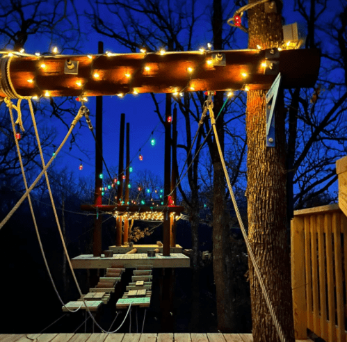 A wooden structure with colorful lights at dusk, surrounded by trees, creating a cozy outdoor atmosphere.