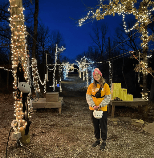 A person stands on a lit path surrounded by trees adorned with string lights, wearing a helmet and smiling.