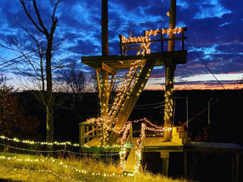 A wooden structure adorned with string lights against a twilight sky, surrounded by trees and a scenic view.