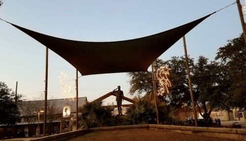 A large shade sail covers a statue, surrounded by trees and festive lights, under a clear evening sky.