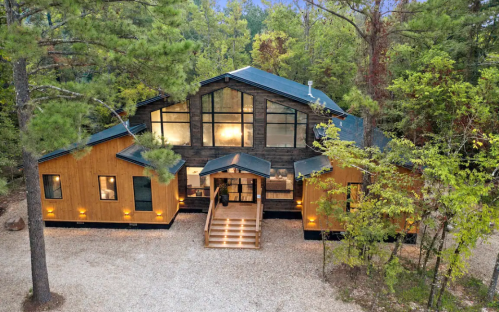 A modern wooden house surrounded by trees, featuring large windows and a welcoming entrance.