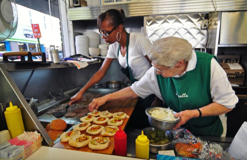 Two women prepare food in a busy kitchen, one grilling meat and the other assembling sandwiches with toppings.