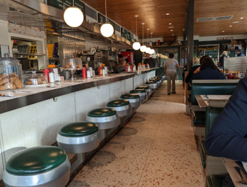 A retro diner interior with a long counter, green stools, and patrons enjoying their meals. Bright, inviting atmosphere.