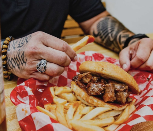 A close-up of a hand with tattoos holding a fry above a BBQ sandwich and fries on a red-checkered paper.