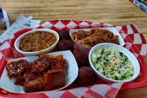 A red tray with barbecue meat, coleslaw, baked beans, pulled pork, and fried balls, all served with checkered paper.