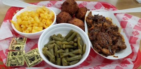 A tray with macaroni and cheese, green beans, pulled pork, and cornbread balls, accompanied by butter packets.