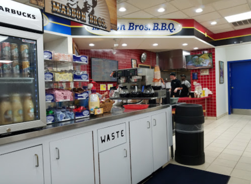 Interior of a BBQ restaurant with a counter, drinks display, and staff preparing food in the background.