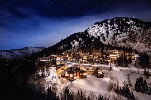 A snowy mountain landscape at night, with illuminated houses nestled among trees and hills under a starry sky.