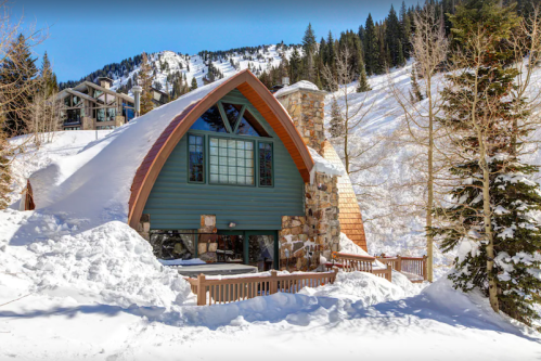 A cozy A-frame cabin surrounded by snow-covered trees and mountains on a sunny winter day.