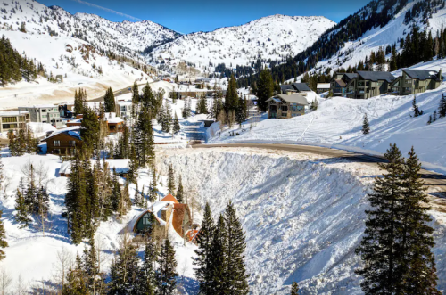A snowy landscape with mountains, trees, and modern homes, showcasing a large snowbank near a road.