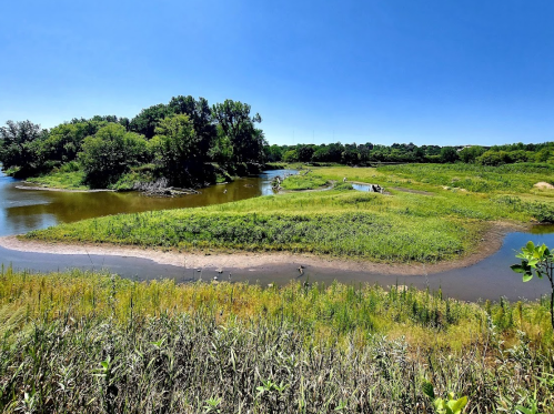 A serene landscape featuring a winding river, lush green grass, and trees under a clear blue sky.