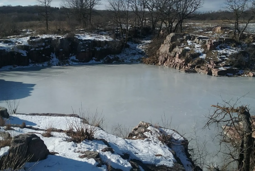 A frozen pond surrounded by rocky terrain and sparse trees, with patches of snow on the ground.