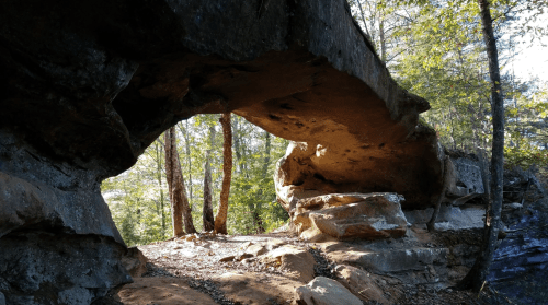 A natural rock arch surrounded by trees and sunlight, with a rocky path beneath it.