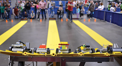 A lineup of LEGO cars on a yellow track, with spectators watching in the background at a robotics event.