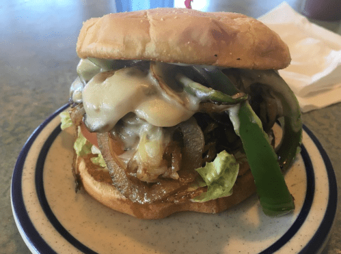A hearty sandwich with grilled onions, green peppers, melted cheese, lettuce, and tomato on a toasted bun.