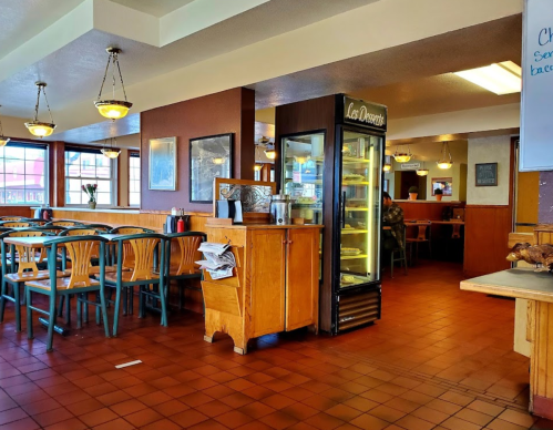A cozy restaurant interior with wooden tables, chairs, and a glass display fridge. Warm lighting and a welcoming atmosphere.