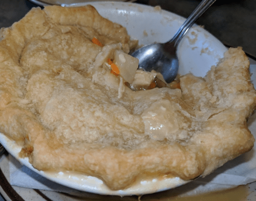 A partially eaten chicken pot pie in a white dish, with flaky crust and visible filling of chicken and vegetables.