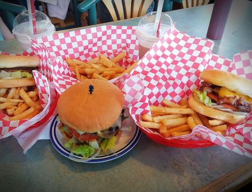 Three baskets of burgers with fries, featuring red and white checkered paper, on a table with milkshakes in the background.