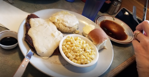 A plate of country-style meal featuring biscuits, corn, gravy-covered meat, and a roll, with a side of syrup.
