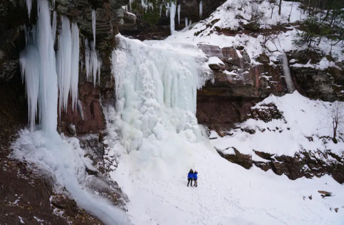 A frozen waterfall surrounded by snow, with two people standing at the base, admiring the icy landscape.