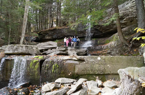 A group of people stands near a rocky waterfall in a forested area, surrounded by trees and natural scenery.