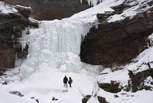 Two hikers stand in front of a frozen waterfall surrounded by snow-covered rocks and trees.