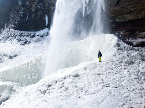 A person in yellow pants stands on a snowy ledge near a frozen waterfall, surrounded by ice and rock formations.