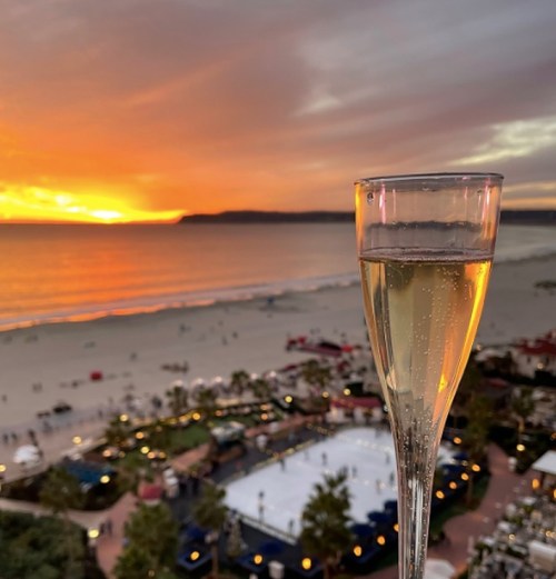 A glass of champagne in the foreground with a sunset over the ocean and beach in the background.