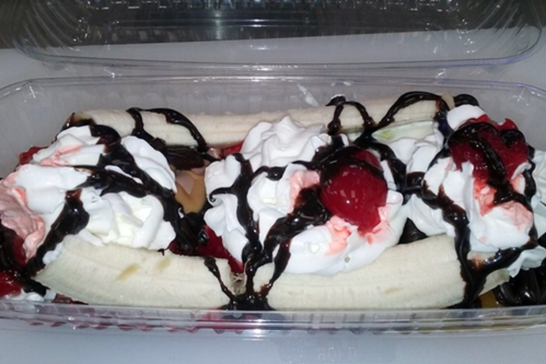 A dessert with bananas, whipped cream, chocolate syrup, and cherries in a clear container.