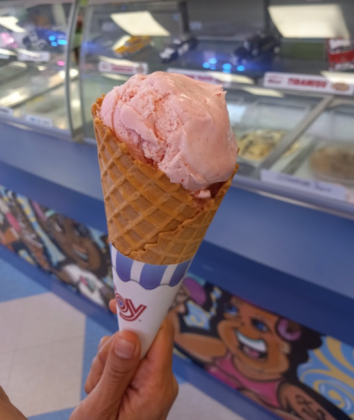 A hand holding a waffle cone filled with pink ice cream, with an ice cream shop display in the background.